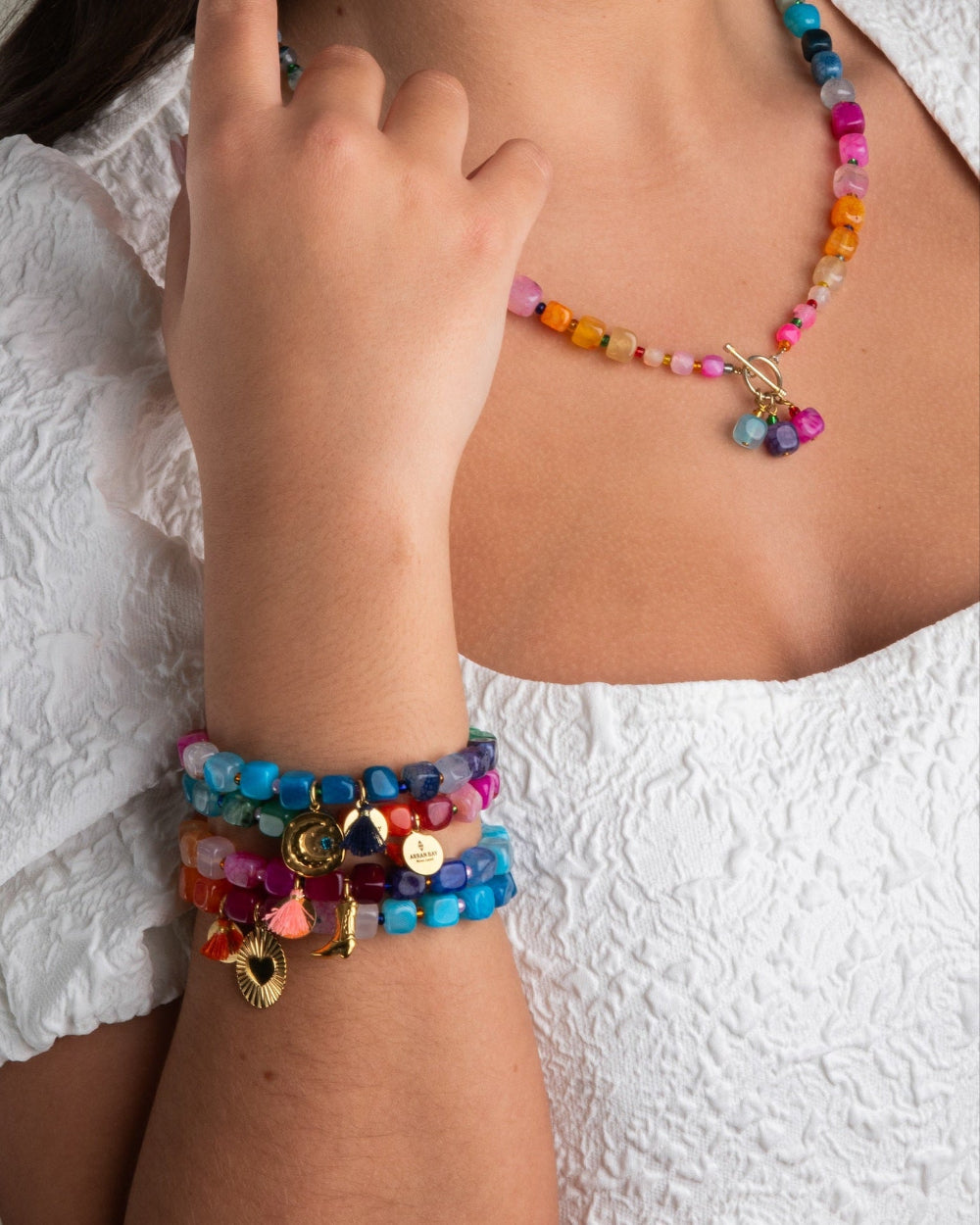 A person wearing a white lace top adorned with colorful beaded bracelets and a matching necklace with agate charms and a gold plate toggle clasp.