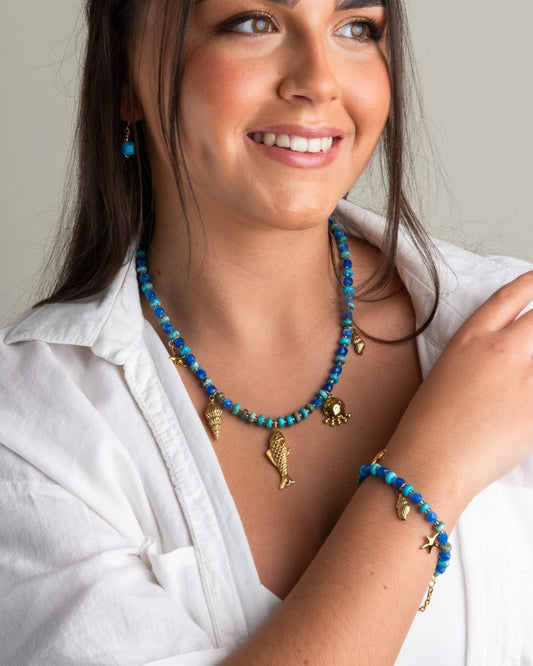 White shirt, showcasing a blue beaded gemstone necklace with gold-plated ocean-themed charms and a matching bracelet.