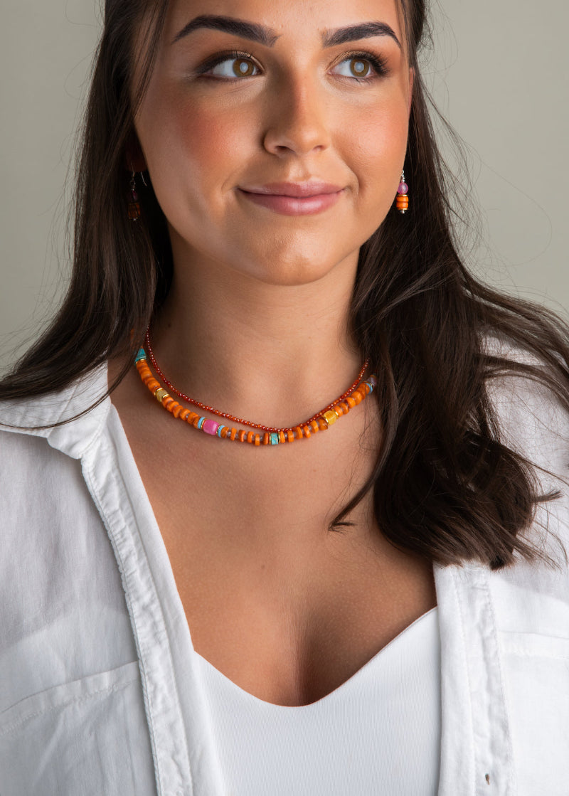 A person wearing an orange shell necklace with gold hematite accents and a white shirt.