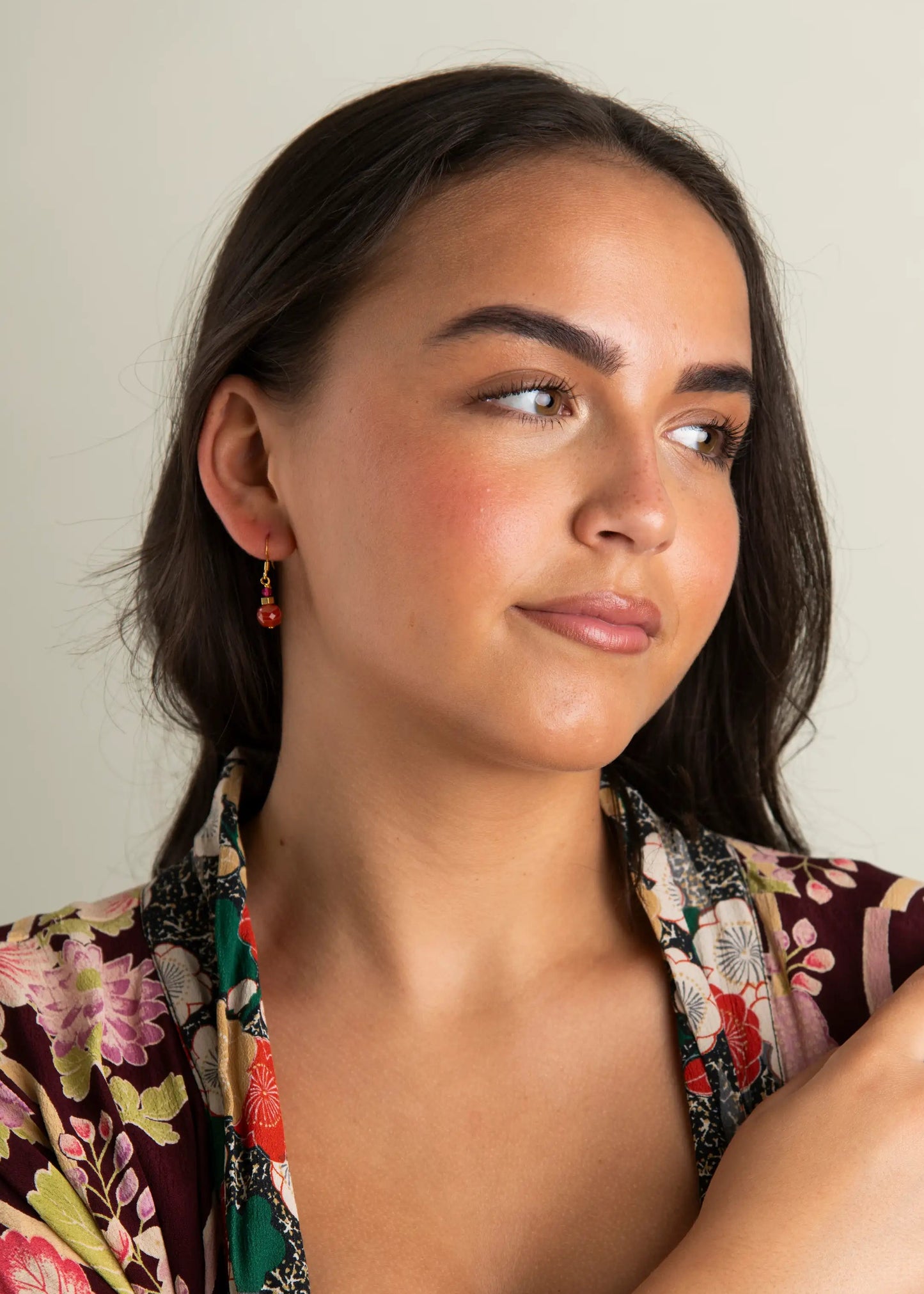A woman wearing a bohemian-style floral print robe in burgundy, green, and pink, accessorised with a beaded bracelet and small rust orange gemstone earrings, set against a plain light grey background.