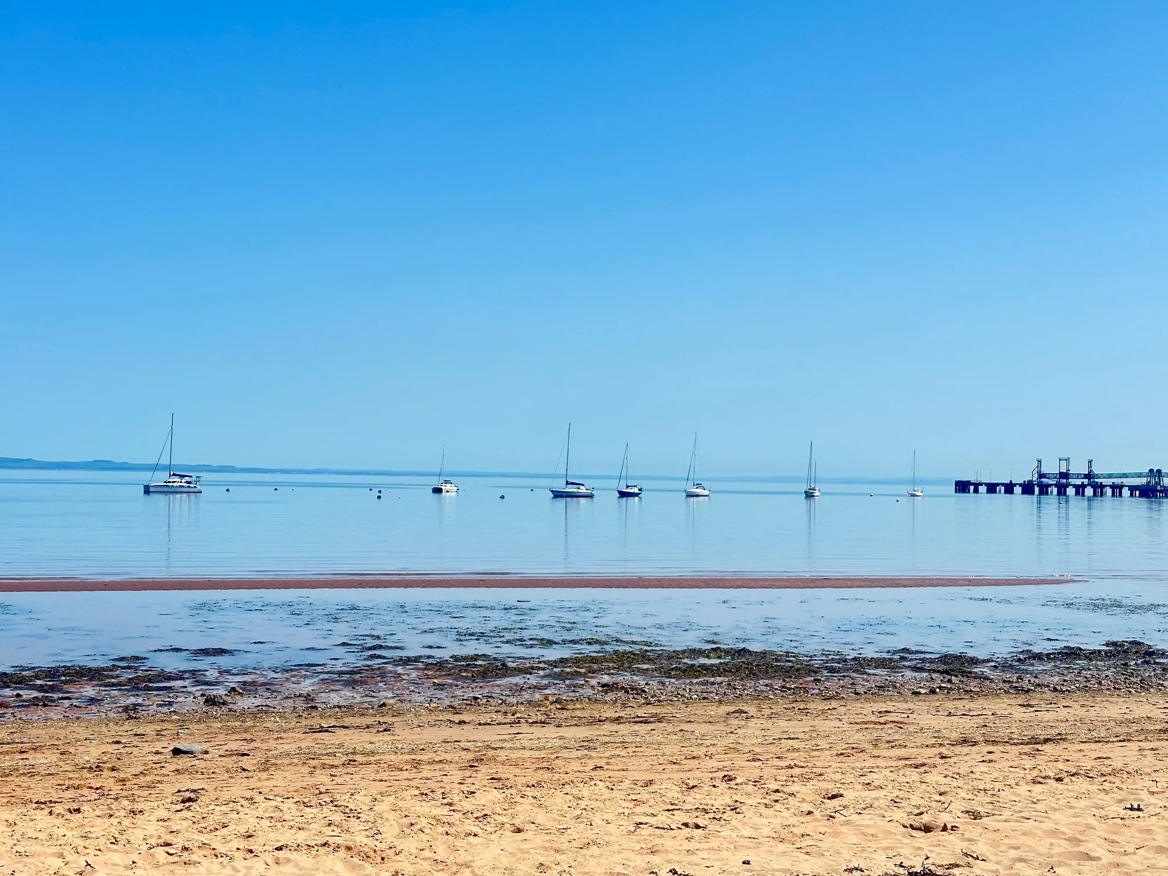 A scenic coastal landscape with a sandy beach in the foreground, calm blue water, several sailboats anchored in the distance, and a pier on the right under a clear blue sky.