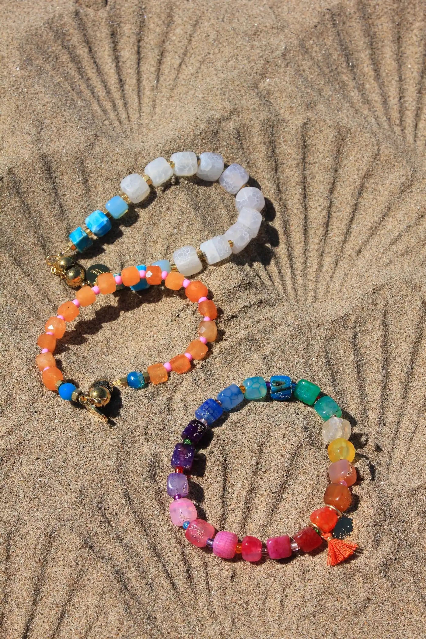Three colorful beaded bracelets arranged on sand. One bracelet is made of white translucent beads with blue and gold accents, one is orange and blue with gold accents, and one is a rainbow gradient with small tassel details.