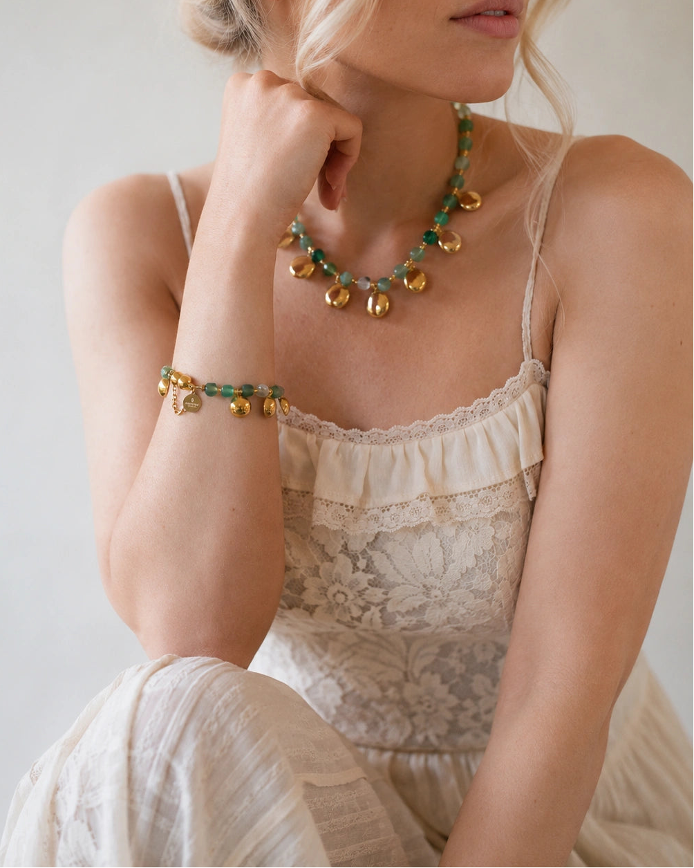 A woman wearing a bohemian-style gold and green beaded necklace and matching bracelet, styled with a cream lace camisole top against a neutral light gray background.