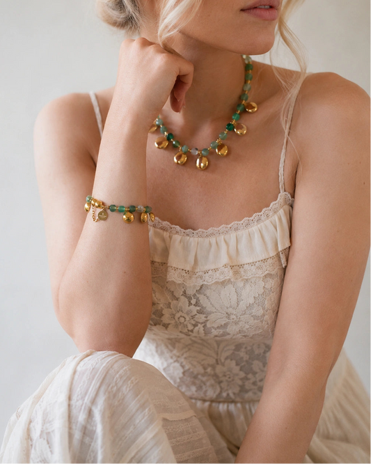 A woman wearing a bohemian-style gold and green beaded necklace and matching bracelet, styled with a cream lace camisole top against a neutral light gray background.